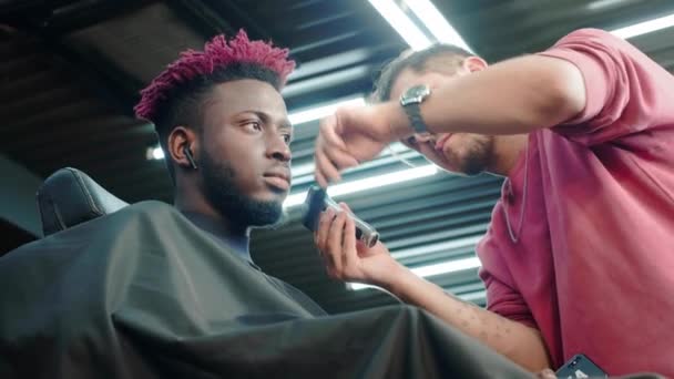 Närbild: en frisör rakar av sig håret på huvudet på en afroamerikan. Gör en snygg frisyr, använder en elektrisk rakapparat. Dreadlocks är röda på huvudet. — Stockvideo