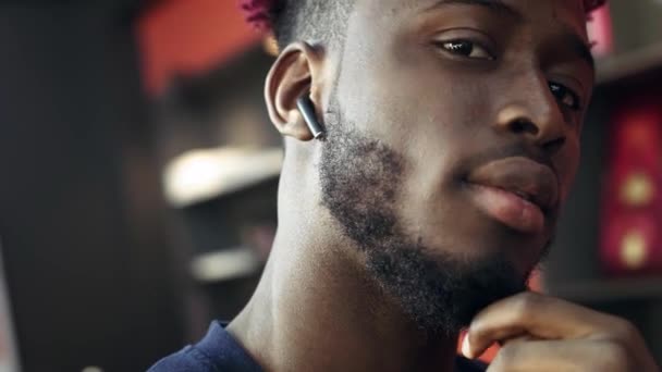 Retrato: Hombre afroamericano con barba mira a la cámara. Utiliza auriculares inalámbricos de moda, escucha música. Nuevas tecnologías inalámbricas . — Vídeos de Stock