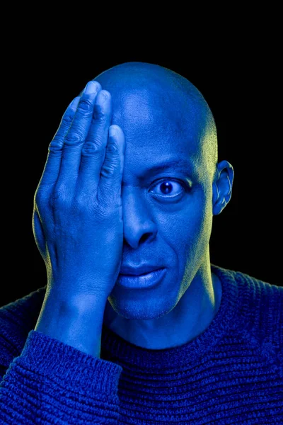 Tek gözü eli eli kapalı, siyahi bir adam ve şaşkınlık ifadesi. Stüdyo fotoğrafı, siyah arkaplanda mavi ışık izole edildi. Çaresizlik konsepti. Dikey
