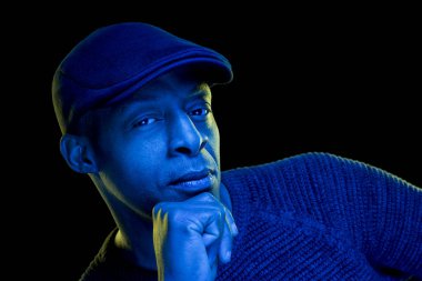 Mavi ve sarı ışıklı siyah bir adam, düz bir şapka takıyor, siyah arka planda izole edilmiş, kameraya bakıyor. Düşünceli bir kavram. Kopyalama uzayı ile yatay