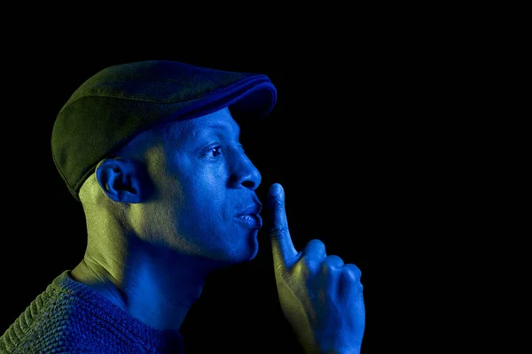 Mavi ve sarı ışığı olan siyah bir adam, parmakları dudaklarında sessizlik hareketi yapıyor, düz bir şapka takıyor, yana bakıyor. Siyah arka planda izole edilmiş, sessizlik kavramı. Kopyalama uzayı ile yatay