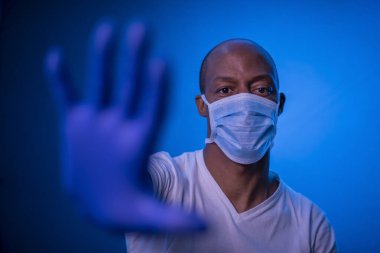 Coronavirus 'u durdur, Afrikalı Amerikalı adam koronavirüsü durdurma işaretini gösterir. Stüdyoda mavi arka planda koruyucu maske ve eldiven takar. Yüzüne odaklan. Covid-19 konsepti.