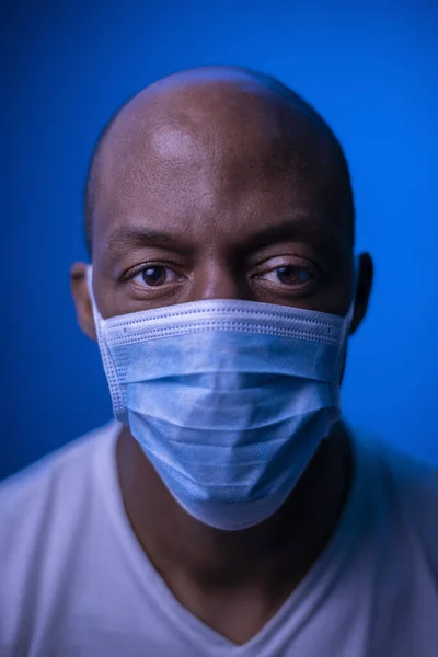 Coronavirus, beyaz gömlekli ve koruyucu maskeli Afro-Amerikan bir adamın resmi, kameraya bakıyor. Covid-19 konsepti. Mavi arkaplanda stüdyo çekimi.