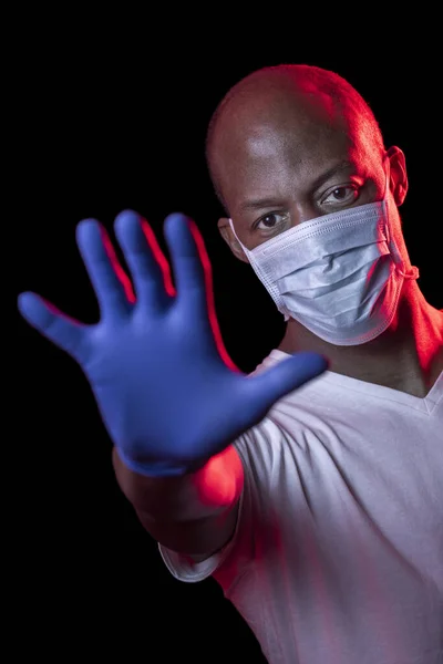 Coronavirus 'u durdur, Afrikalı Amerikalı adam koronavirüsü durdurma işaretini gösterir, koruyucu maske takar ve stüdyonun karanlık arka planında eldivenler takar. Yüzüne odaklan. Covid-19 konsepti.