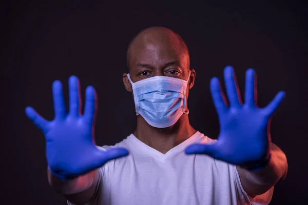 DUR Coronavirus, Afro-Amerikalı adam iki eliyle koronavirüsü durdurma işaretini gösteriyor, koruyucu bir maske takıyor ve arka planda eldiven takıyor. Yüzüne odaklan. COVID-19 kavramı.