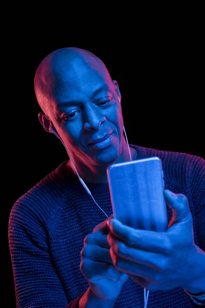 Siyah arka planda izole edilmiş akıllı telefon ve kulaklıklı siyah bir adamın mavi ve kırmızı ışıklarıyla portresi. Mutluluk ifadesi.