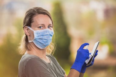 Coronavirus konsepti, koruyucu tıbbi maskeli ve ameliyat eldivenli sarışın kadın akıllı telefonuna antiseptik jel sürüyor. Salgın önleme COVID-19