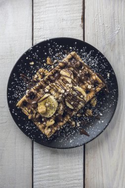 Muz dilimleri, yulaf, ceviz ve çikolatayla kaplı Belçika waffle 'ı beyaz bir ahşap masanın üzerinde siyah bir tabakta servis edilir. Üst görünüm.