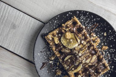 Muz dilimleri, yulaf, ceviz ve çikolata ile kaplı waffle, beyaz ahşap bir masanın üzerinde siyah bir tabakta servis edilir. Kopyalama alanı ile üst görünümü kapat.