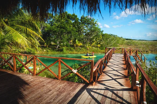 Dock along seven colored lagoon with canoe passing by surrounded by tropical plants in Bacalar, Quintana Roo, Mexico