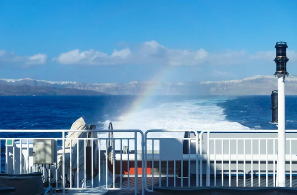 Back of ferry boat with rainbow from splashing water with blurred view on Santorini cliffs, Greece