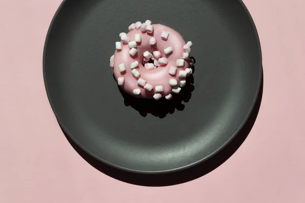 Açık mercan zemininde koyu gri seramik tabakta beyaz marşmelovlu pembe bir donat. Geometrik güneşli, gölgeli yaratıcı kompozisyon. Kutlama günü için sanat fikri