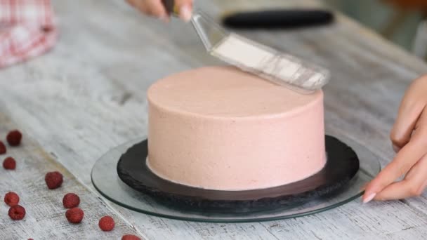 Hands of female confectionery chef using pastry scraper and rotating cake stand to decorate handmade cake with pink cream frosting in kitchen. — Stock Video