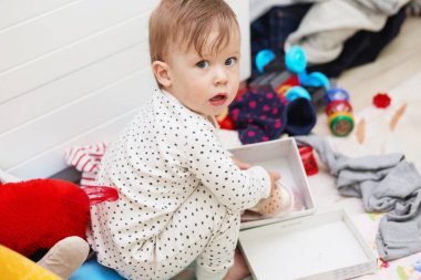 Şirin bebek kız yere çömelme pijama ayakkabılarını dağınık şeyler ve oyuncaklar arasında koymak çalışır