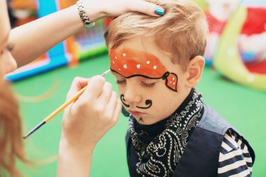 Master making aqua makeup on boys face. Halloween party. A little boy in a pirate costume and a makeup on his face is having a good time at the Halloween party. Face painting kids. clipart