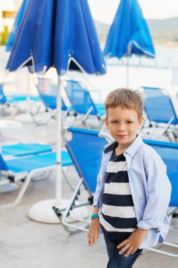 Mavi şemsiye ve şezlonglar arasında küçük bir çocuk duruyor 