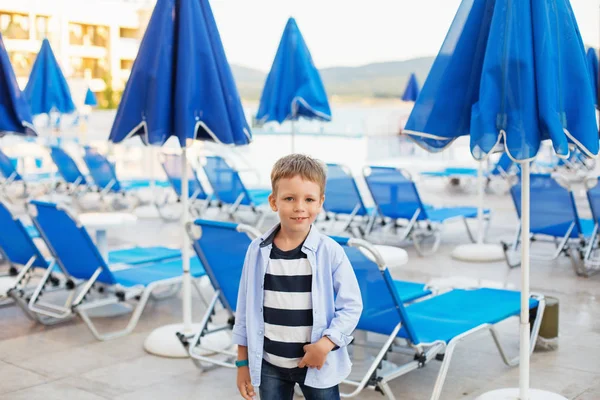 A small boy stands among the blue umbrellas and sun loungers on — Stock Photo, Image