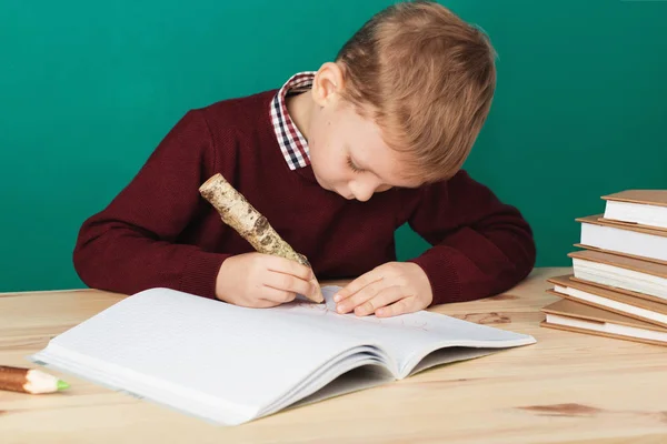 Smiling little schoolboy writing notes in class against green wa — Stock Photo, Image