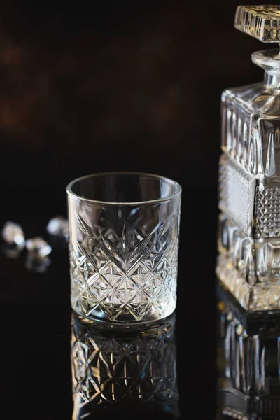 Empty Glass for whiskey or bourbon with and a crystal square decanter on the black reflective surface background