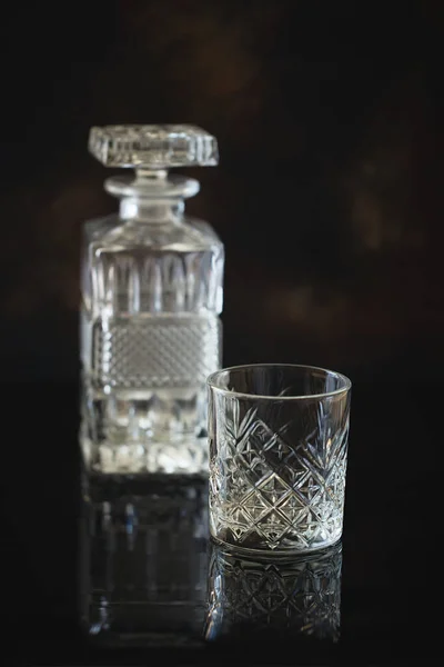 Empty Glass for whiskey or bourbon with and a crystal square decanter on the black reflective surface background