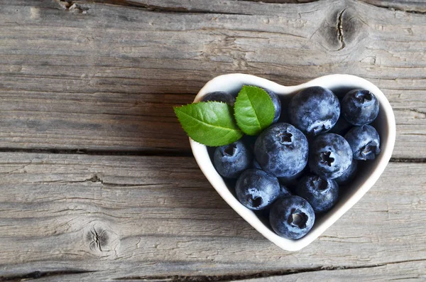 Verse biologische bosbessen in een wit hart vormige kom op een jute doek op rustieke houten tafel. Blueberry.Bilberries.Healthy eten, veganistische voeding, dieet en voeding concept. — Stockfoto