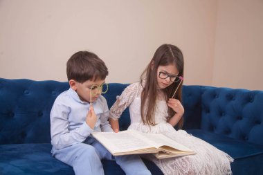 brother and sister are sitting on the sofa in paper glasses and playfully reading a book