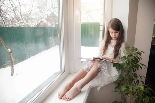 Beyaz elbiseli genç bir kız pencere kenarında oturur ve bir kitap okur.