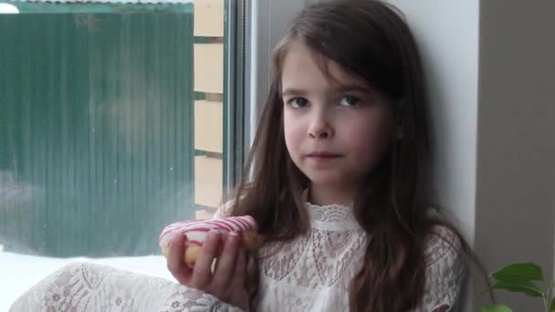 女の子は大きな窓に座って甘いドーナツを食べています — ストック動画