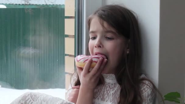 女の子は大きな窓に座って甘いドーナツを食べています — ストック動画