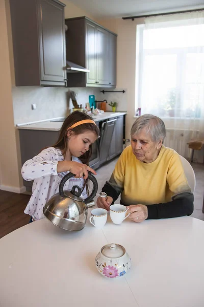 Büyükanne ve torun mutfaktaki beyaz masada oturup çay içerler.