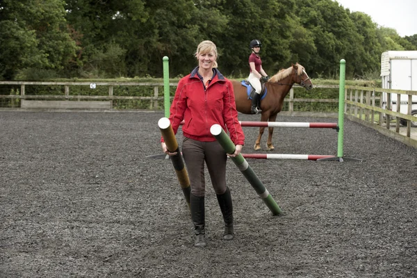 Chica montando un pony e instructor llevando postes de valla — Foto de Stock