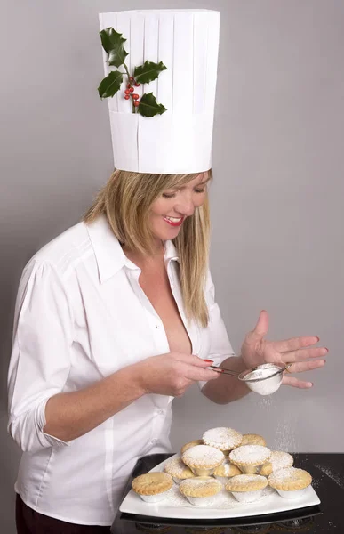 Woman wearing a disposable chef's hat sprinkles icing sugar onto mince pies Stock Picture