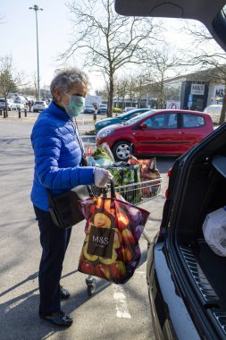 Southampton, İngiltere, İngiltere. Mart 2020. Coronavirus salgını sırasında korunmak için maske ve lastik eldiven takan yaşlı bir kadın market alışverişini arabaya yüklüyor..