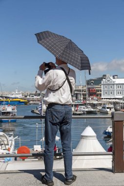 Cape Town, Güney Afrika. 2019. Güneş gölgesi olarak şemsiye tutan bir adam ve rıhtımın fotoğrafını çekmek için kamera kullanan bir adam..