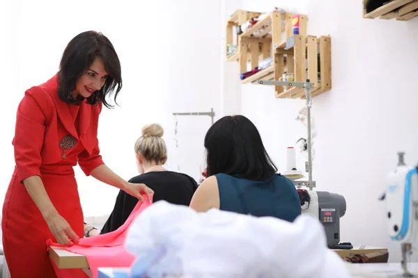 Schöne Mädchen in einem roten Kleid in einem Schneideratelier. Exklusive Maßanfertigung von Kleidern auf Bestellung. Das Mädchen näht ein Kleid an einer Nähmaschine. Europäerin arbeitet in Nähwerkstatt. — Stockfoto