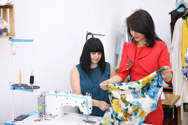 Hermosa chica en un vestido rojo en un estudio de sastrería. Exclusiva confección de vestidos a pedido. La chica cose un vestido en una máquina de coser. Mujer europea trabaja en un taller de costura . —  Fotos de Stock