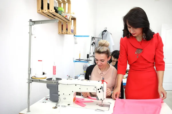 Beautiful girl in a red dress in a tailoring studio. Exclusive tailoring of dresses to order. The girl sews a dress on a sewing machine. European woman works in a sewing workshop.