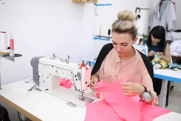 テーラリングスタジオで赤いドレスを着た美しい女の子。注文するドレスの排他的な仕立て。少女はミシンでドレスを見た。ヨーロッパの女性は縫製工場で働く. — ストック写真
