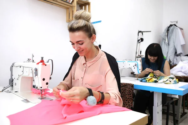 Hermosa chica en un vestido rojo en un estudio de sastrería. Exclusiva confección de vestidos a pedido. La chica cose un vestido en una máquina de coser. Mujer europea trabaja en un taller de costura . —  Fotos de Stock