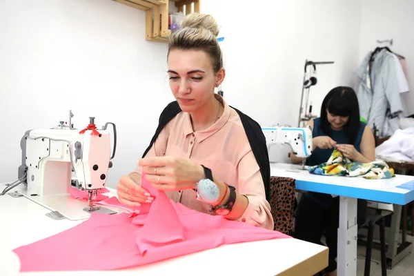 Hermosa chica en un vestido rojo en un estudio de sastrería. Exclusiva confección de vestidos a pedido. La chica cose un vestido en una máquina de coser. Mujer europea trabaja en un taller de costura . —  Fotos de Stock