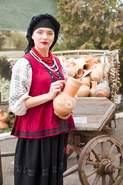 Hermosa chica en vestido nacional. Llevando una olla antigua en sus manos. Ropa antigua de finales del siglo XIX. Hermoso vestido y falda en una mujer. El concepto de vida rural, tradiciones nacionales —  Fotos de Stock