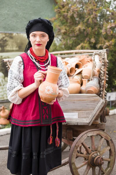 Bella ragazza in abito nazionale. Teneva in mano un'antica pentola. Abbigliamento antico della fine del XIX secolo. Bellissimo vestito e gonna su una donna. Il concetto di vita rurale, tradizioni nazionali — Foto Stock