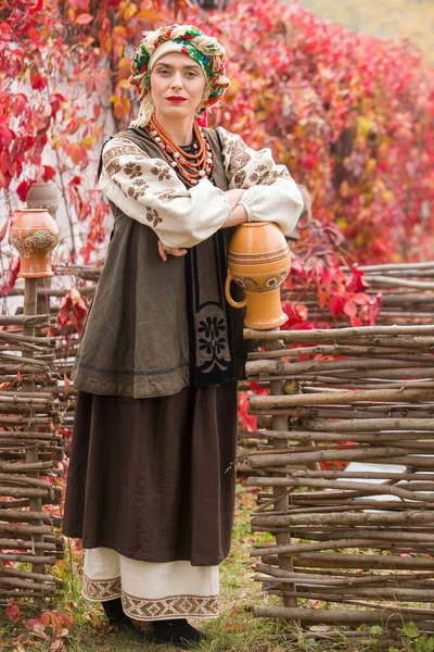 穿着民族服装的漂亮姑娘。用一个古老的陶罐。19世纪末的古董服装。女人穿上漂亮的衣服和裙子.美丽的秋天和树叶。十九世纪末的衣服 — 图库照片