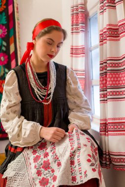 19. yüzyılın ulusal kostümü içinde güzel bir genç kız. Pencerenin yanında güzel bir desen işliyor. Çelenk ve kurdeleyle. Fotoğraftaki köy, kırsal bölge. Başörtüsüyle.
