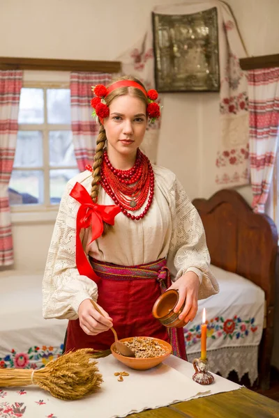 Menina bonita em vestido nacional está preparando um jantar festivo. Em uma grinalda bonita e um vestido bordado vermelho. Celebração familiar e celebração do costume nacional. Tigela com kutia - tradicional — Fotografia de Stock