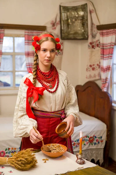 Belle fille en robe nationale prépare un dîner festif. Dans une belle couronne et une robe rouge brodée. Célébration familiale et célébration des coutumes nationales. Bol avec kutia - traditionnel — Photo