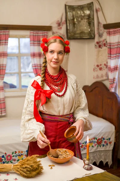 Όμορφο κορίτσι με εθνικό φόρεμα ετοιμάζει ένα εορταστικό δείπνο. Με ένα όμορφο στεφάνι και ένα κόκκινο κεντημένο φόρεμα. Οικογενειακή γιορτή και εορτασμός των εθνικών εθίμων. Μπολ με kutia - παραδοσιακό — Φωτογραφία Αρχείου