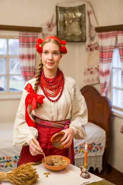 Menina bonita em vestido nacional está preparando um jantar festivo. Em uma grinalda bonita e um vestido bordado vermelho. Celebração familiar e celebração do costume nacional. Tigela com kutia - tradicional — Fotografia de Stock