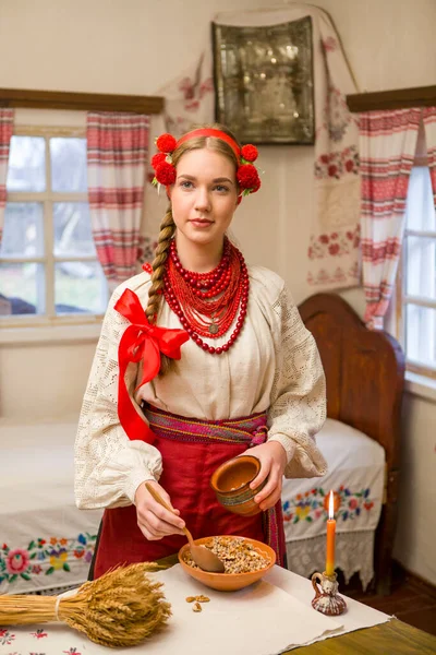 Menina bonita em vestido nacional está preparando um jantar festivo. Em uma grinalda bonita e um vestido bordado vermelho. Celebração familiar e celebração do costume nacional. Tigela com kutia - tradicional — Fotografia de Stock