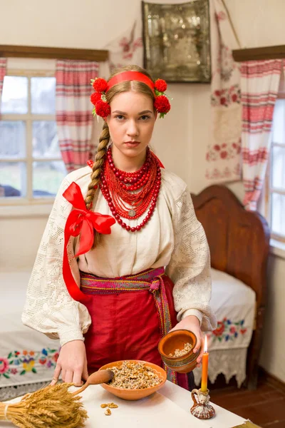 Gyönyörű lány nemzeti ruhában ünnepi vacsorát készít. Egy gyönyörű koszorúban és egy piros hímzett ruhában. Családi ünnepség és a nemzeti szokás ünneplése. Bowl kutia - hagyományos — Stock Fotó